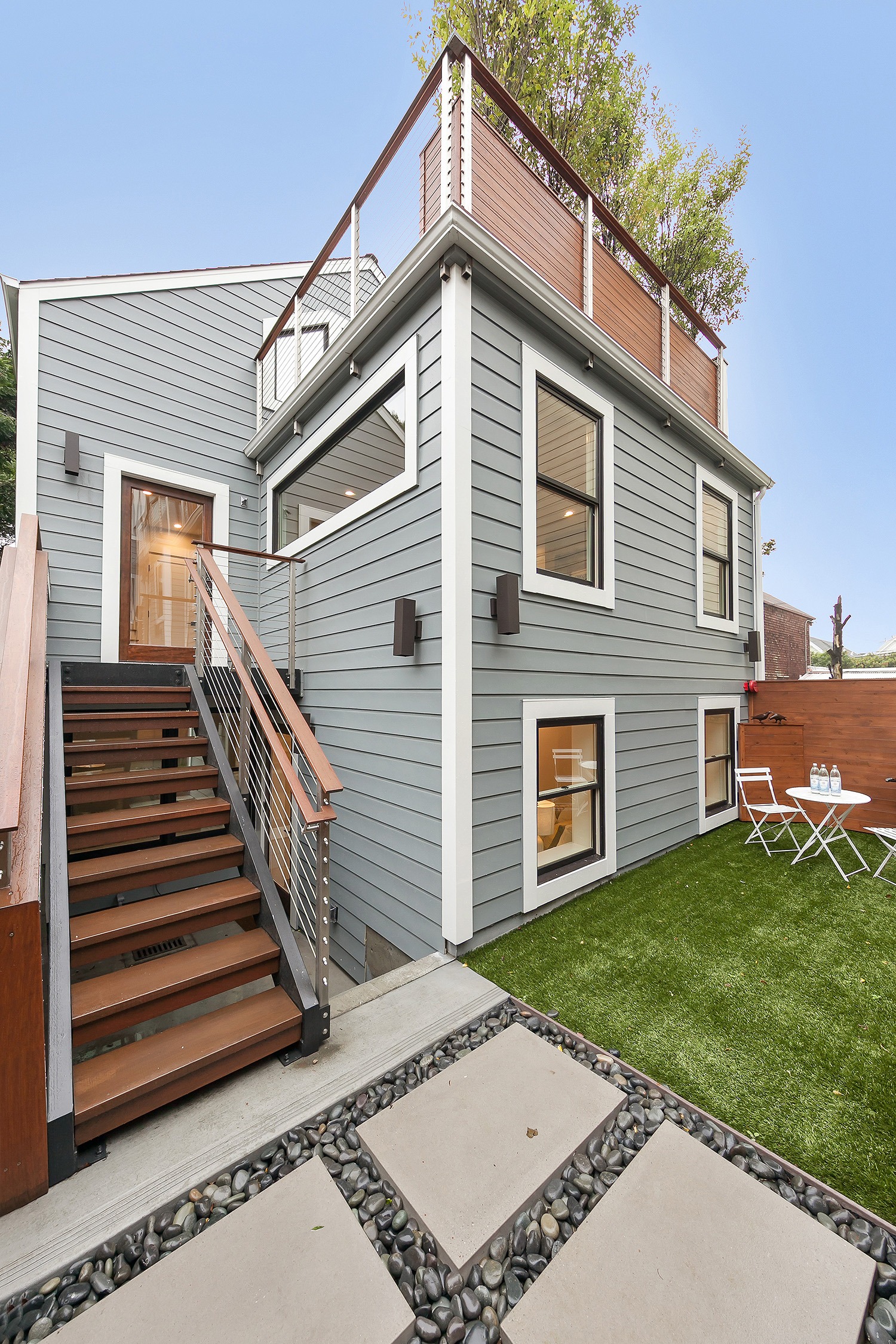 Modern two-story gray house with a wooden deck, stairs leading up, and a small outdoor seating area on artificial grass. Ideal for outdoor relaxation.