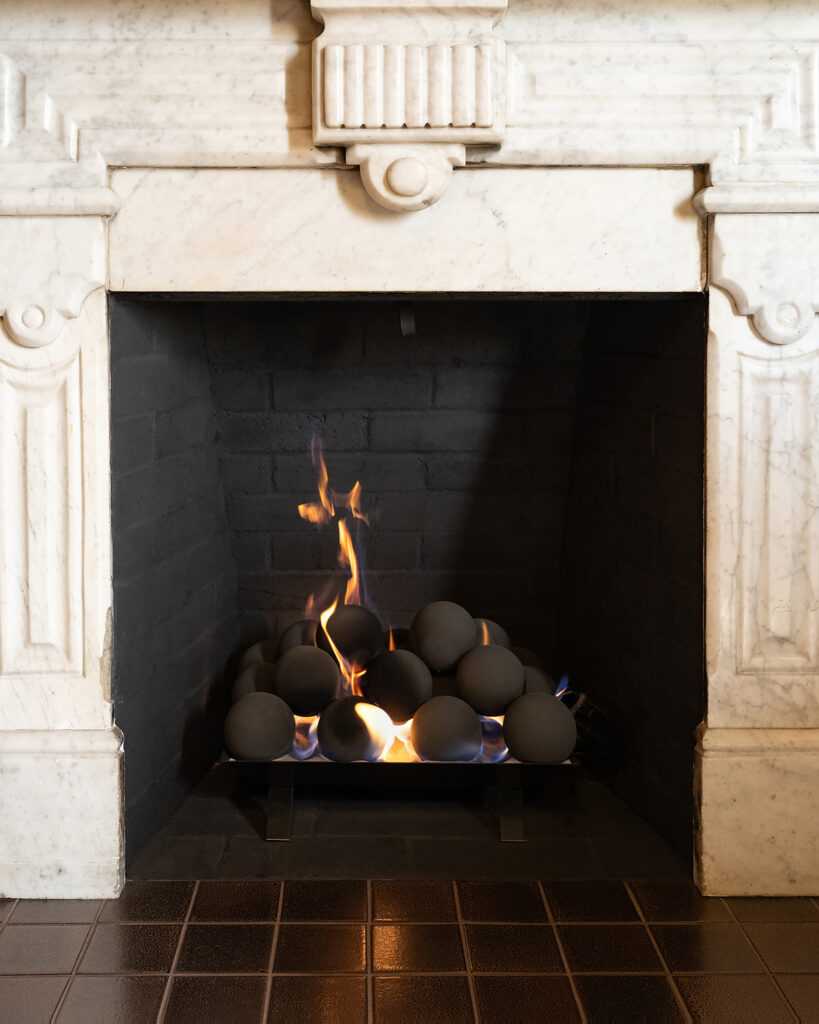 A modern fireplace features a vibrant flame above a bed of smooth gray decorative stones. Elegant marble surrounds enhance the cozy atmosphere.