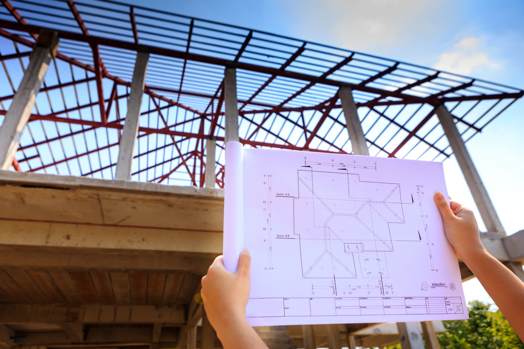 Blueprint held in hands shows construction plans, with a skeletal building structure in the background under a clear blue sky.