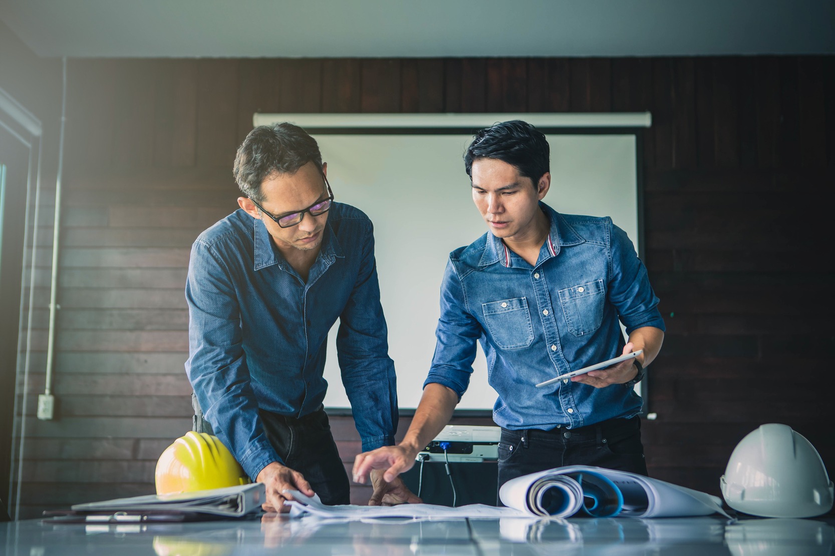 Two engineers discuss project plans on a table, analyzing documents and a tablet. Safety helmets are in the foreground, indicating a construction context.